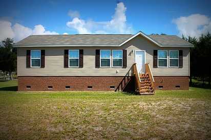 A welcoming single-story home with brick foundation sits on spacious grounds, featuring bright blue shutters and a wooden front porch staircase.
