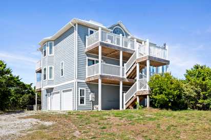 A spacious three-story coastal property with multiple balconies and an elevated design, surrounded by natural greenery and open land under clear blue skies.